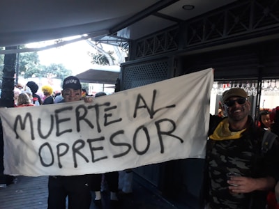 A group of people, some in costumes, are holding a large banner with the text 'MUERTE AL OPRESOR'. The scene appears to be set in an outdoor area with trees visible in the background and a covered walkway. One individual is wearing a cap labeled 'SWAT' and another person wearing sunglasses and a bandana is standing nearby. The mood is festive and possibly part of a protest or celebration.