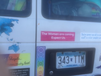 The image shows the back of a vehicle with multiple stickers and a Minnesota license plate (343 TTM). Some notable stickers include a pink one that reads 'The Women are coming. Expect Us.' and other stickers with various messages and colorful designs. The inside of the vehicle appears cluttered with different items visible through the window.