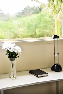 A serene indoor setting features a white vase with white flowers placed on a countertop. Beside it lies a closed black book labeled 'Holy Bible'. A sleek black desk lamp stands on the opposite end. The background reveals a window covered with raindrops, overlooking a lush green landscape.