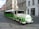 A green and white urban tram-like vehicle is parked on a cobblestone street in a European city. The vehicle has 'URBAN' written on a round front panel and is designed for promoting environmentally friendly transport with the slogan 'Choose green'. Historic buildings with ornate facades are seen in the background, one of which is pink with intricate architectural details. The street is quiet with few people or vehicles.