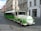 A green and white urban tram-like vehicle is parked on a cobblestone street in a European city. The vehicle has 'URBAN' written on a round front panel and is designed for promoting environmentally friendly transport with the slogan 'Choose green'. Historic buildings with ornate facades are seen in the background, one of which is pink with intricate architectural details. The street is quiet with few people or vehicles.