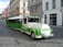 A green and white urban tram-like vehicle is parked on a cobblestone street in a European city. The vehicle has 'URBAN' written on a round front panel and is designed for promoting environmentally friendly transport with the slogan 'Choose green'. Historic buildings with ornate facades are seen in the background, one of which is pink with intricate architectural details. The street is quiet with few people or vehicles.