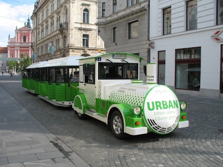 A green and white urban tram-like vehicle is parked on a cobblestone street in a European city. The vehicle has 'URBAN' written on a round front panel and is designed for promoting environmentally friendly transport with the slogan 'Choose green'. Historic buildings with ornate facades are seen in the background, one of which is pink with intricate architectural details. The street is quiet with few people or vehicles.