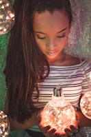 A young girl with long hair gazes downwards, holding a large, glowing light bulb filled with string lights. She wears a striped off-the-shoulder top and stands against a shimmery, textured background. The warm glow of the lights creates a soft, dreamy atmosphere.
