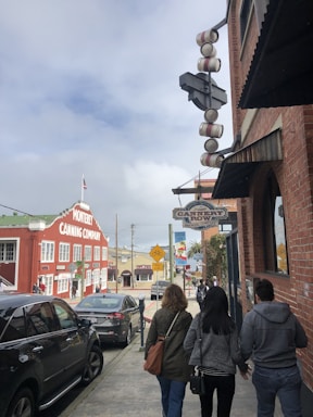 A street scene features several people walking along the sidewalk beside a brick building. Signs for Cannery Row Brewing Company and Monterey Canning Company are prominently displayed. Cars are parked along the street, and a cloudy sky is visible in the background.