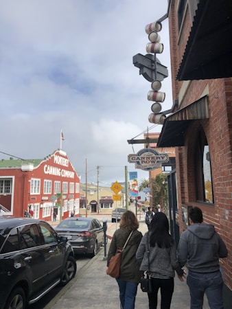 A street scene features several people walking along the sidewalk beside a brick building. Signs for Cannery Row Brewing Company and Monterey Canning Company are prominently displayed. Cars are parked along the street, and a cloudy sky is visible in the background.
