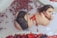 A person is lounging in a milky bath adorned with red rose petals and cinnamon sticks. The individual is wearing black lace lingerie with a red ribbon tied around the waist. The mood is serene and intimate, with soft lighting enhancing the calm atmosphere.