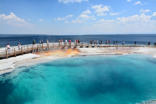 A group of people walk along a wooden boardwalk next to a vibrant geothermal pool. The pool displays striking blue and turquoise hues, contrasted by the orange and white deposits around its edges. In the background, a vast body of water extends to the horizon under a partly cloudy sky.