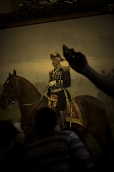 A portrait painting depicts a military officer in ornate uniform, sitting confidently on a brown horse. The officer wears a hat and has multiple medals on his chest. The painting is being observed by a crowd, whose silhouettes are partially visible in the foreground. An arm is raised, possibly taking a photo or pointing at the artwork.