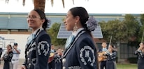 A group of musicians, dressed in traditional mariachi outfits, are performing outdoors. Two women are prominently in the foreground, wearing dark blue suits adorned with intricate white embroidery and large flowers in their hair. In the background, other musicians can be seen playing instruments such as the guitar and trumpet. There are solar panels and trees present, set against a building.