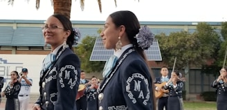 A group of musicians, dressed in traditional mariachi outfits, are performing outdoors. Two women are prominently in the foreground, wearing dark blue suits adorned with intricate white embroidery and large flowers in their hair. In the background, other musicians can be seen playing instruments such as the guitar and trumpet. There are solar panels and trees present, set against a building.