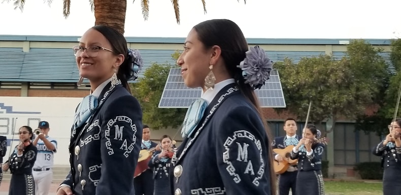 A group of musicians, dressed in traditional mariachi outfits, are performing outdoors. Two women are prominently in the foreground, wearing dark blue suits adorned with intricate white embroidery and large flowers in their hair. In the background, other musicians can be seen playing instruments such as the guitar and trumpet. There are solar panels and trees present, set against a building.