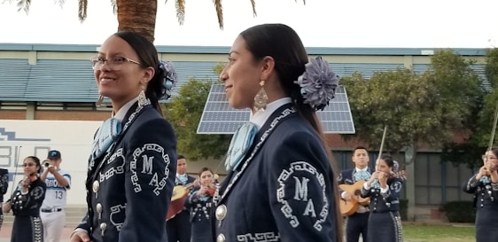 A group of musicians, dressed in traditional mariachi outfits, are performing outdoors. Two women are prominently in the foreground, wearing dark blue suits adorned with intricate white embroidery and large flowers in their hair. In the background, other musicians can be seen playing instruments such as the guitar and trumpet. There are solar panels and trees present, set against a building.