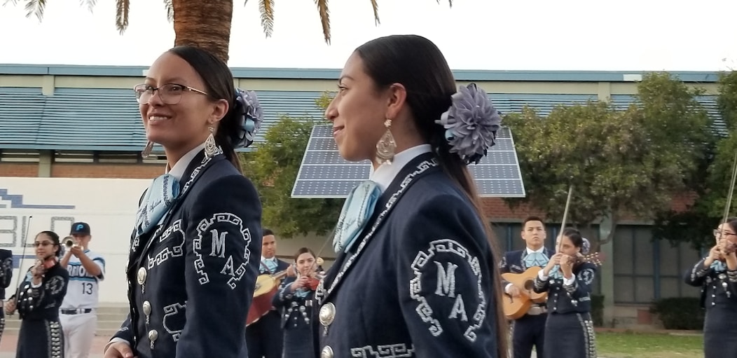 A group of musicians, dressed in traditional mariachi outfits, are performing outdoors. Two women are prominently in the foreground, wearing dark blue suits adorned with intricate white embroidery and large flowers in their hair. In the background, other musicians can be seen playing instruments such as the guitar and trumpet. There are solar panels and trees present, set against a building.