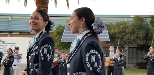 A group of musicians, dressed in traditional mariachi outfits, are performing outdoors. Two women are prominently in the foreground, wearing dark blue suits adorned with intricate white embroidery and large flowers in their hair. In the background, other musicians can be seen playing instruments such as the guitar and trumpet. There are solar panels and trees present, set against a building.