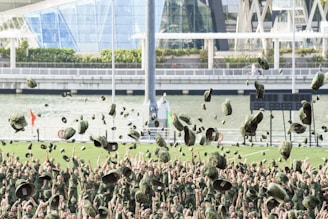 A large group of people in military uniforms are celebrating by throwing their hats into the air. The background features a river, modern architectural structures, and greenery.