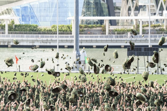 A large group of people in military uniforms are celebrating by throwing their hats into the air. The background features a river, modern architectural structures, and greenery.