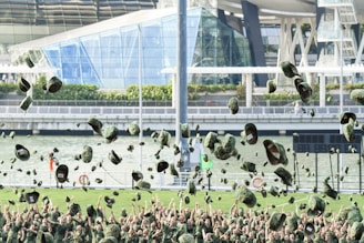A group of people in military uniforms are celebrating by throwing their camouflage caps into the air. In the background, there is a modern building with glass structures near a waterfront. The scene is lively and filled with joyful expressions.