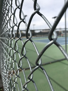 A close-up view of a metal chain-link fence with a blurred background of outdoor tennis courts and equipment in the distance. The focus is on the intertwined wires of the fence, highlighting its texture and structure.