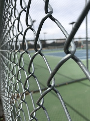 A close-up view of a metal chain-link fence with a blurred background of outdoor tennis courts and equipment in the distance. The focus is on the intertwined wires of the fence, highlighting its texture and structure.