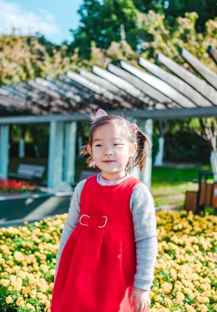 A young child wearing a bright red dress stands in a garden with vibrant yellow flowers. The child has a cheerful expression, with hair tied in pigtails adorned with pink accessories. In the background, there's a wooden pergola and lush green trees under a clear blue sky.