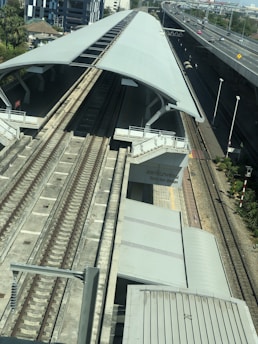 A modern elevated train station with two sets of railway tracks running side by side. The station is covered with a large, curved metal roof. Adjacent to the station is a highway with multiple lanes and sparse traffic, including several cars. Surrounding the infrastructure are some trees and buildings, indicating an urban setting.