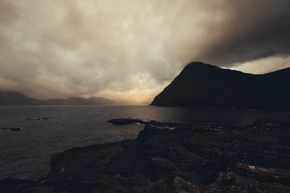 calm body of water near mountain during gloomy skies