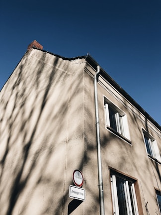 A tall, beige stucco building with visible wear and shadow patterns on its surface. It has modern windows and a red-trimmed street sign attached near its base, reading 'Anlieger frei' in German. The sky is clear and intensely blue, contrasting with the muted tones of the building.