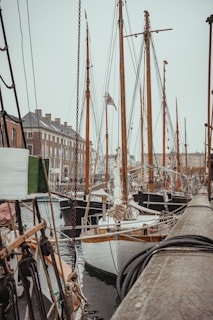 A harbor scene featuring several wooden sailboats with tall masts docked alongside a wooden pier. The background shows a line of brick buildings typical of a historic port city. The rigging and ropes of the boats are prominently visible, adding texture and complexity to the scene.