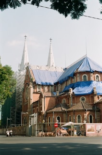 A large, historic church with two tall white spires topped with crosses is under renovation. The building features red brick walls and sections covered with blue tarps. Scaffolding is visible on one side, and there is a fence around the building with a construction sign attached. A few people on mopeds are present in the foreground.