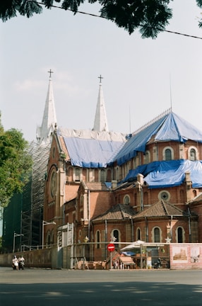 A large, historic church with two tall white spires topped with crosses is under renovation. The building features red brick walls and sections covered with blue tarps. Scaffolding is visible on one side, and there is a fence around the building with a construction sign attached. A few people on mopeds are present in the foreground.