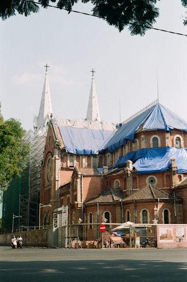 A large, historic church with two tall white spires topped with crosses is under renovation. The building features red brick walls and sections covered with blue tarps. Scaffolding is visible on one side, and there is a fence around the building with a construction sign attached. A few people on mopeds are present in the foreground.