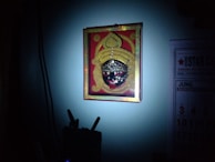 A colorful artwork of a deity or mask with intricate details and bright colors, framed and hung on a wall. The image is predominantly dark with a spotlight focusing on the artwork, creating a dramatic contrast. There's also a glimpse of a calendar on the wall, partially illuminated.