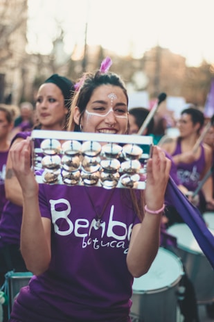 A woman is holding a shiny metallic percussion instrument and smiling. She has decorative face paint and a feather in her hair. She is surrounded by other people, all of whom appear to be part of a musical group or parade, wearing similar purple shirts with a logo on them.
