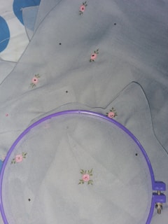 A piece of light-colored fabric with small pink rose patterns and green leaves embroidered on it. A purple embroidery hoop is partially shown, used to hold the fabric taut.