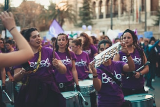 A group of people are gathered, wearing purple shirts with writing on them. They are playing musical instruments like drums and tambourines, seemingly as part of a street performance or march. The crowd appears energetic and focused.