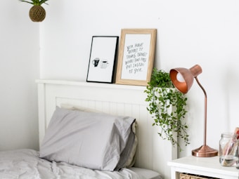 A cozy and neatly arranged bedroom corner features a bed with gray sheets and pillows. Above the white headboard, two framed artworks are displayed; one with a motivational quote and the other with coffee cup illustrations. A copper-colored desk lamp sits on the white bedside table next to glass jars. A small potted plant with trailing leaves adds a touch of greenery, hanging down from the headboard.