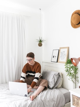 man in brown sweater sitting on bed using laptop computer