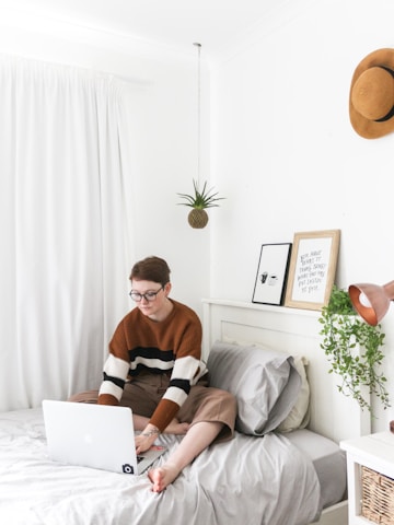 man in brown sweater sitting on bed using laptop computer