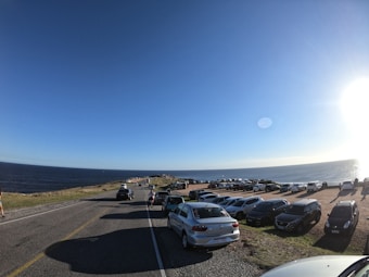 A coastal road with cars parked along the side, overlooking the ocean. The sky is clear with the sun brightly shining, creating reflections on the water. People can be seen walking near the parked cars, and the landscape features grassy areas and a vast expanse of blue sea under a wide sky.