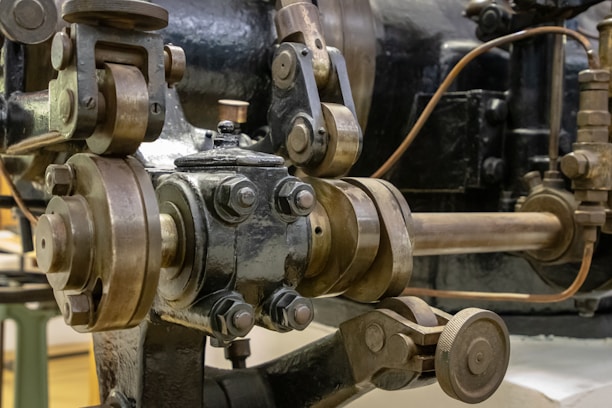 A close-up view of a complex mechanical system with metal gears and levers, featuring a combination of polished and raw metal textures. The intricate components suggest machinery or industrial equipment, possibly part of an engine or other heavy-duty apparatus.