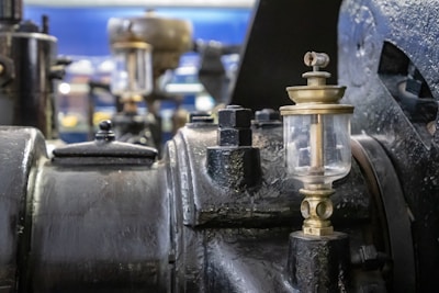 Close-up view of an intricate machine with metal and brass components, including a glass oil feeder prominently featured. The background is blurred, highlighting the mechanical parts and their textures.