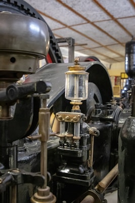 An industrial machine part with metallic components, including a cylindrical container with a glass section. The setting suggests an old factory or industrial environment with visible gears and pipes.