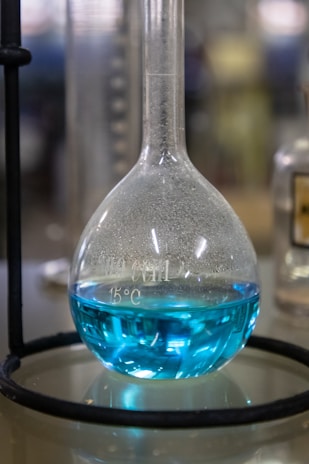 A laboratory flask containing a bright blue liquid is positioned on a reflective surface. The flask has a rounded base with a narrow neck and is held in place by a metal stand. Measurements and temperature markings are visible on the surface of the flask. The background appears blurred, suggesting a lab setting.