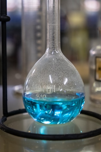 A laboratory flask containing a bright blue liquid is positioned on a reflective surface. The flask has a rounded base with a narrow neck and is held in place by a metal stand. Measurements and temperature markings are visible on the surface of the flask. The background appears blurred, suggesting a lab setting.