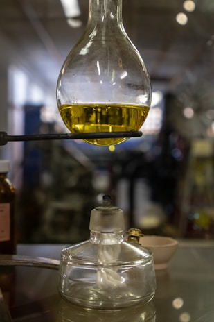 A laboratory setup featuring a round-bottom flask filled with a yellow liquid, supported by a stand. Below it, there is an alcohol burner with a visible wick. The background is blurred, suggesting a laboratory setting.