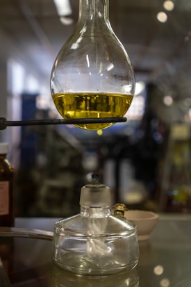 A laboratory setup featuring a round-bottom flask filled with a yellow liquid, supported by a stand. Below it, there is an alcohol burner with a visible wick. The background is blurred, suggesting a laboratory setting.