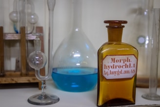 Several laboratory glass containers, including a spherical glass vessel and a brown bottle with a label that reads 'Morph. hydrochl.1 Aq. Amygd. am.49', are arranged on a wooden rack. Another glass container is filled with a blue liquid.