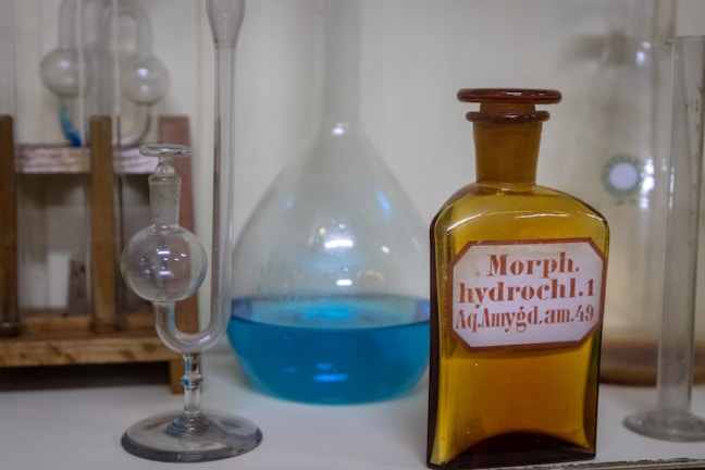 Several laboratory glass containers, including a spherical glass vessel and a brown bottle with a label that reads 'Morph. hydrochl.1 Aq. Amygd. am.49', are arranged on a wooden rack. Another glass container is filled with a blue liquid.