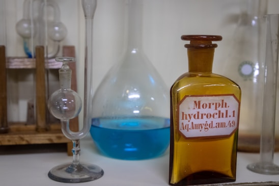 Several laboratory glass containers, including a spherical glass vessel and a brown bottle with a label that reads 'Morph. hydrochl.1 Aq. Amygd. am.49', are arranged on a wooden rack. Another glass container is filled with a blue liquid.